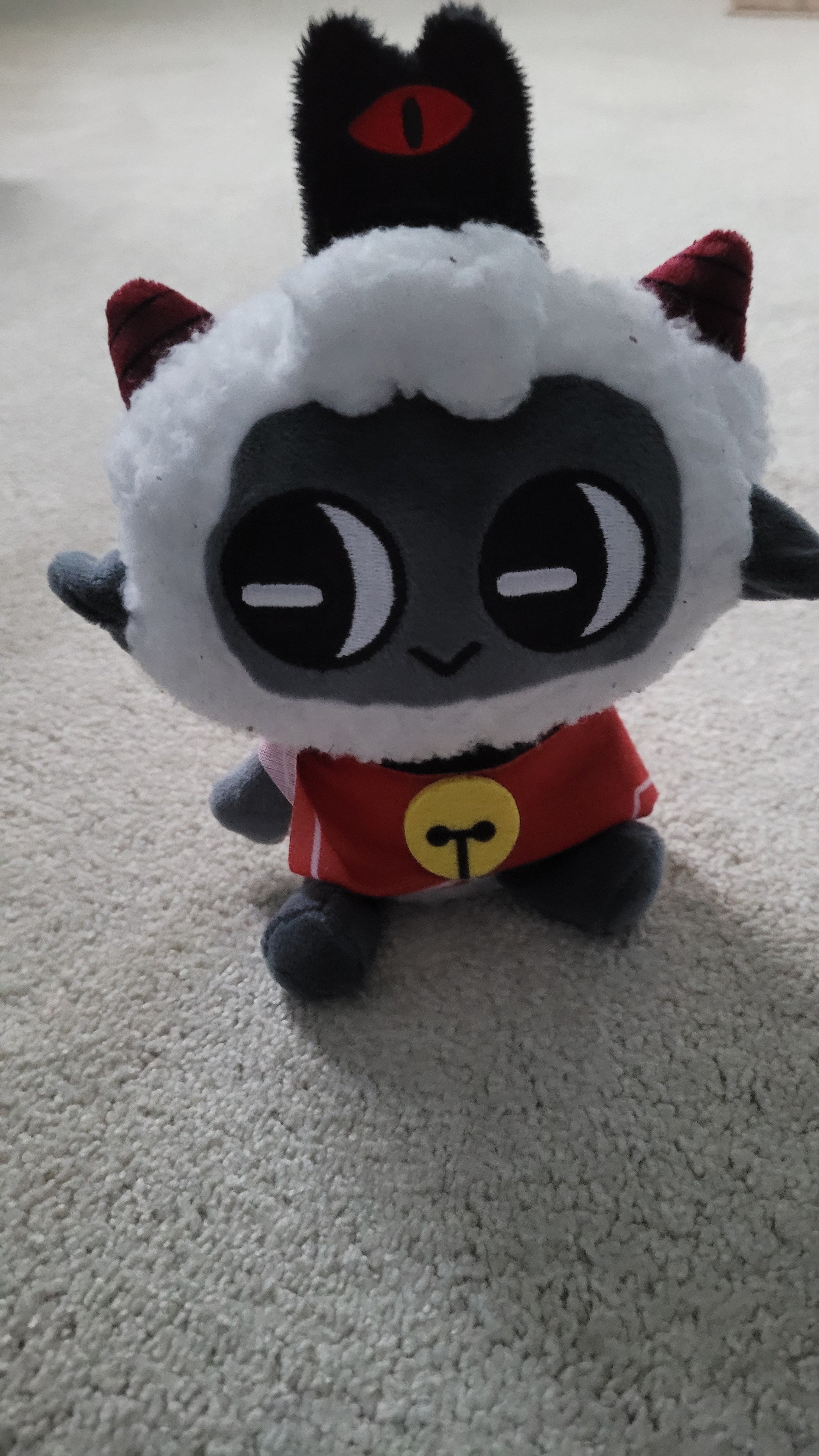 Een plush van het Lam van Cult of the Lamb die zit op zandkleurig tapijt

Het lam zelf is grijskleurig met witte vacht, heeft 2 rode hoorns, draagt een zwarte hoed met een rood oog erop, over z'n schouders een rode cape met zwarte halsband en daaraan een gele bel
