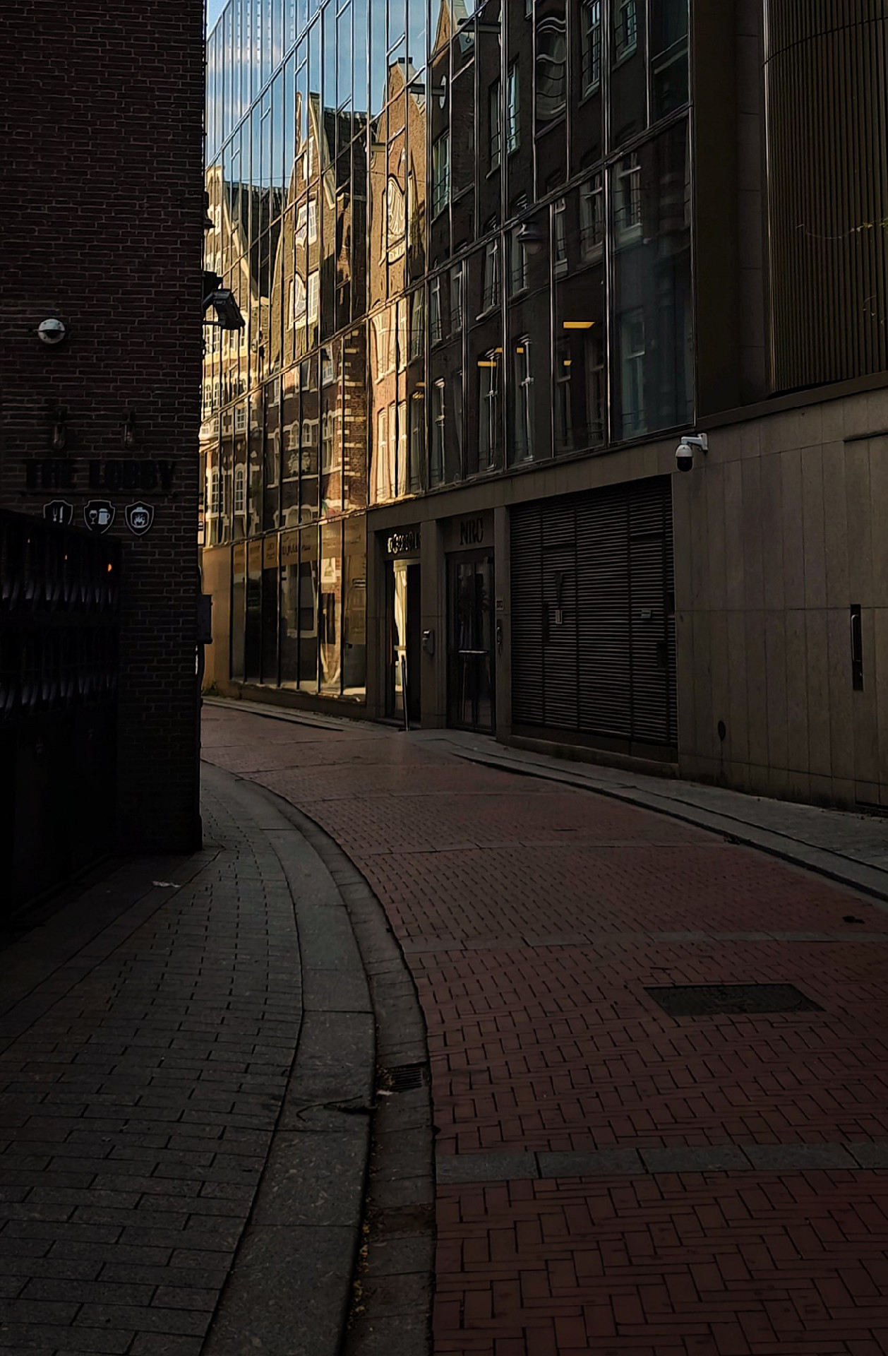 A narrow urban street with brick and stone paving. The scene features a mix of architectural styles including modern concrete structures on the right, and a glass high-rise building visible in the background. Older brick buildings with traditional facades are visible as reflections in the glass facade.