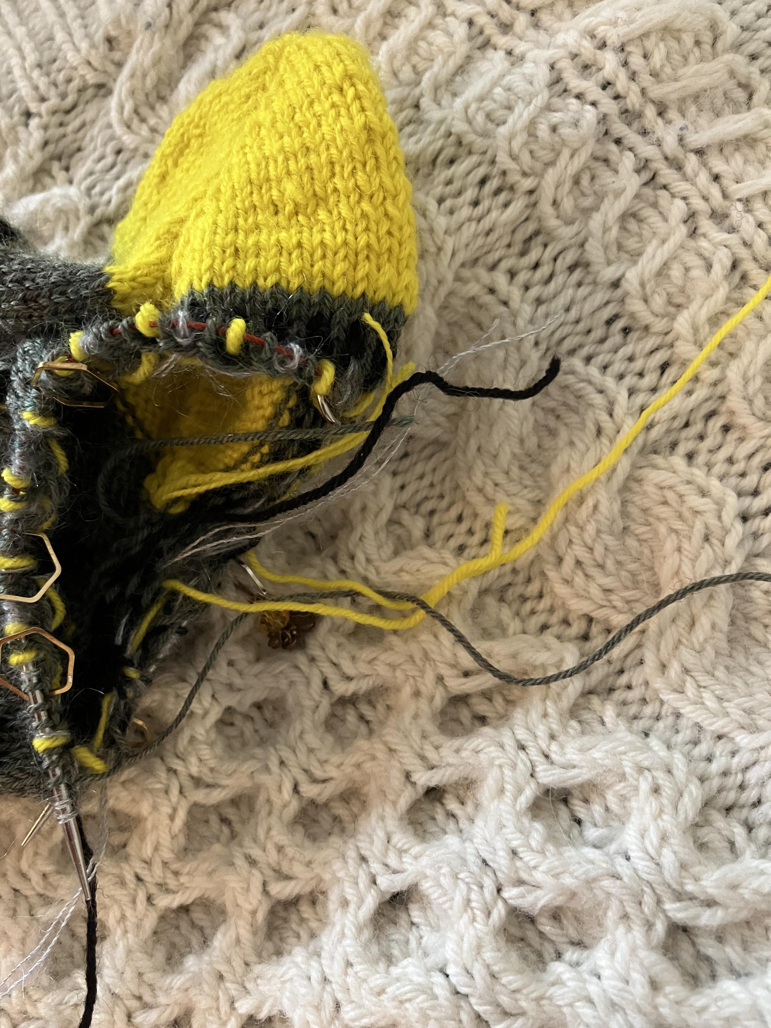 Bright yellow heel sticking up from a partially knit green sock. Yellow, green, black and silver mohair threads are barfing out of the opening inside the smaal circular knitting needle.