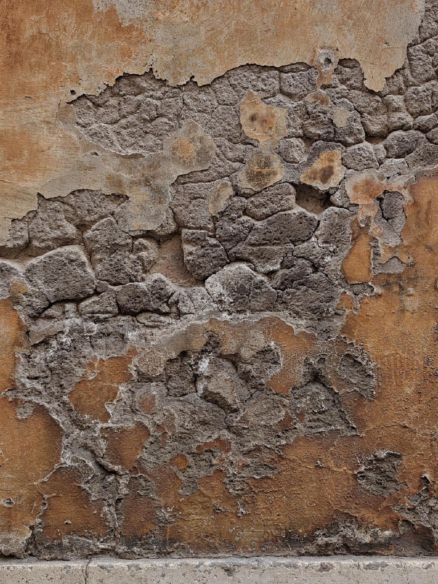 A photo of a section of wall, the yellow-brown plaster has been falling of over a large area revealing cracked layers of gray stone beneath the surface.