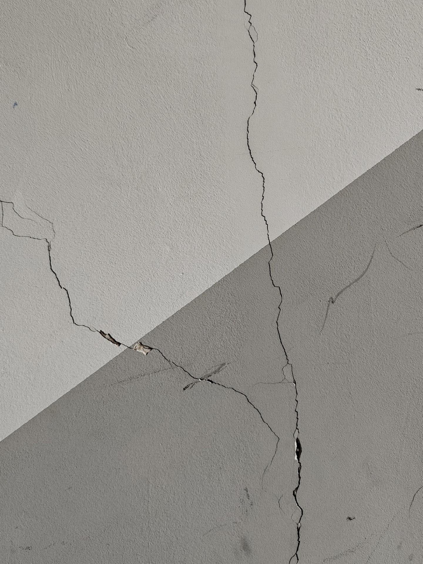 A photo showing a white and grey plastered wall with multiple intersecting cracks running across its surface. The lower right of the wall is grey, the upper left white, the border between the two colours lying diagonally across the image.