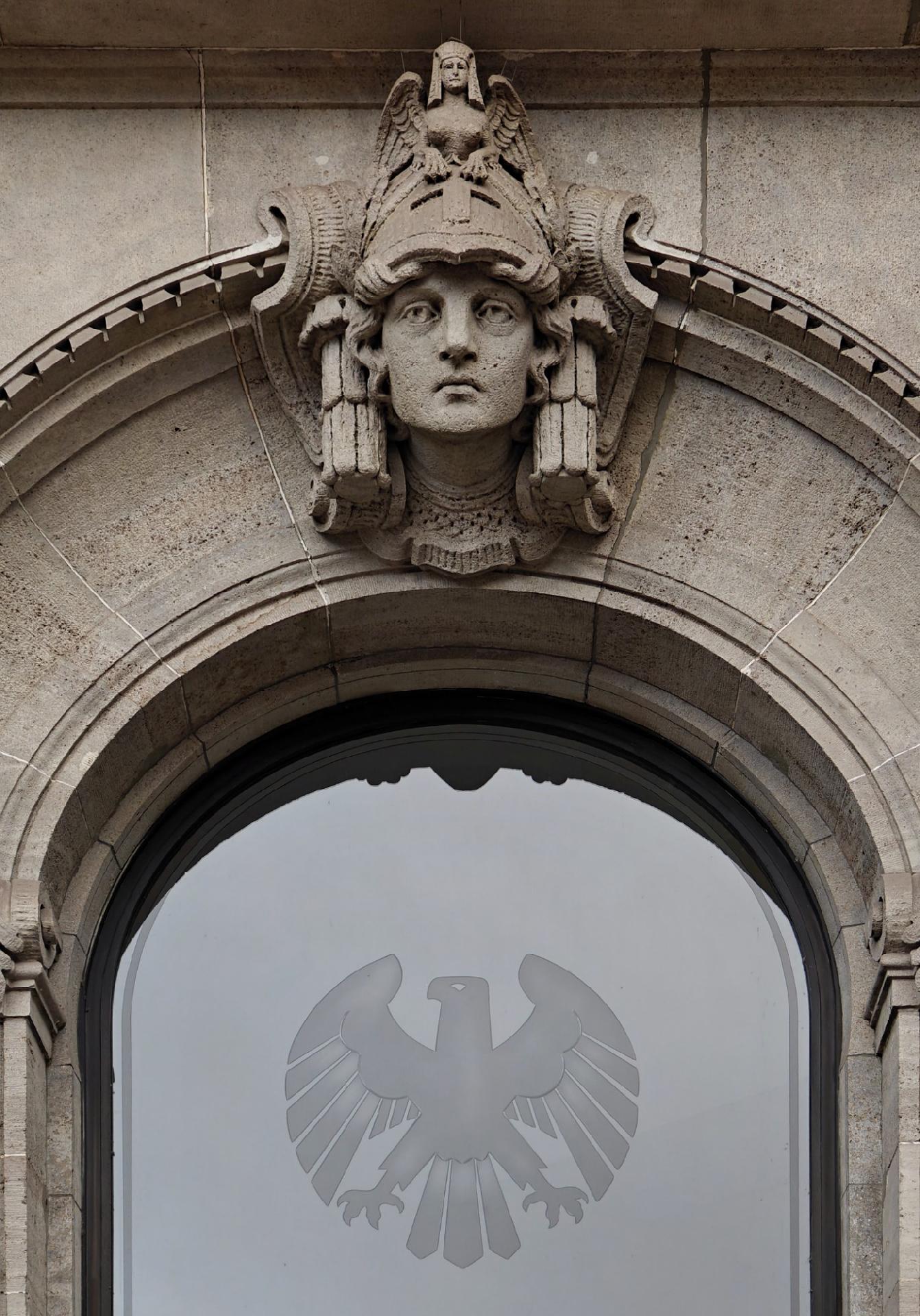 A close up photo of a stone building façade featuring an ornate sculpted head crowned by a small sphinx like creature above an arched window. The window is frosted glass with a stylized eagle.