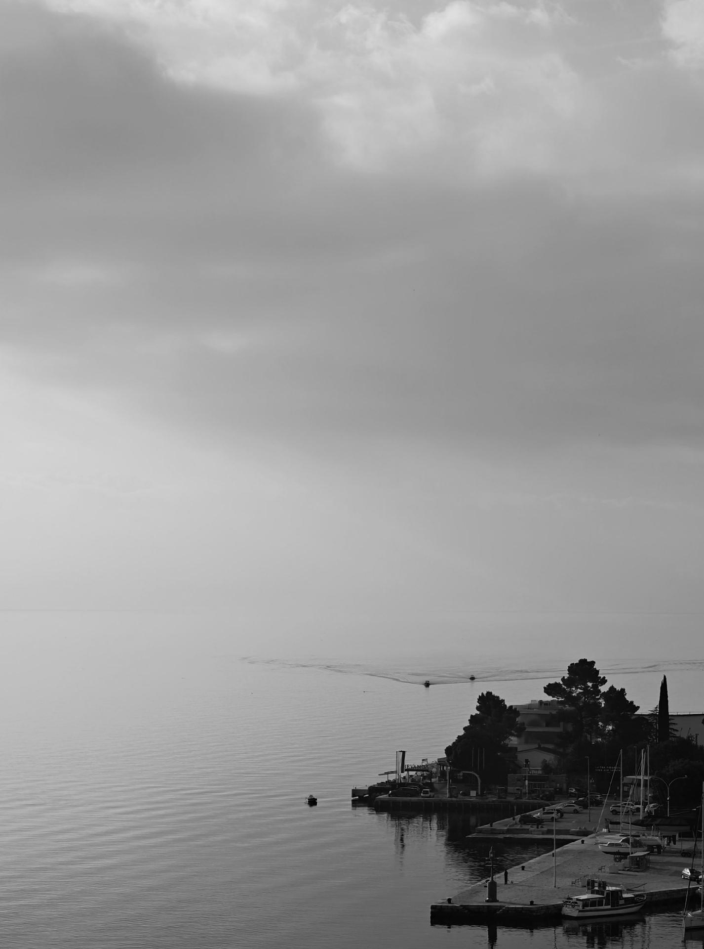 A black and white photo looking down into a coastal marina with calm water. Two small boats coming in from the distance, leaving faint wakes. The horizon is barely visible between the sea and the clody sky.