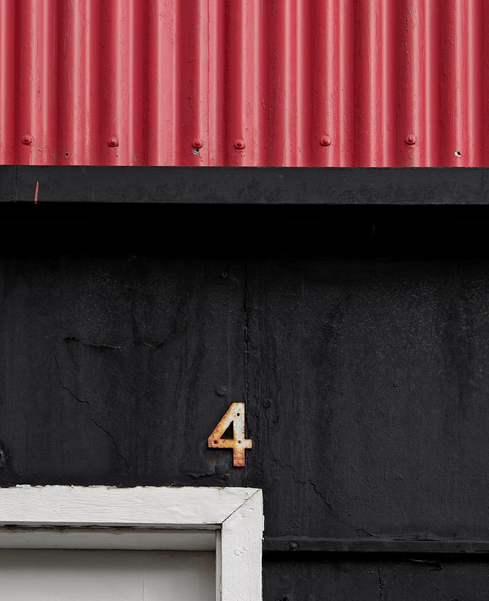 Photo of a side of a house with one part red corrugated metal and another part black. A rusty “4,” hangs over a white door frame.
