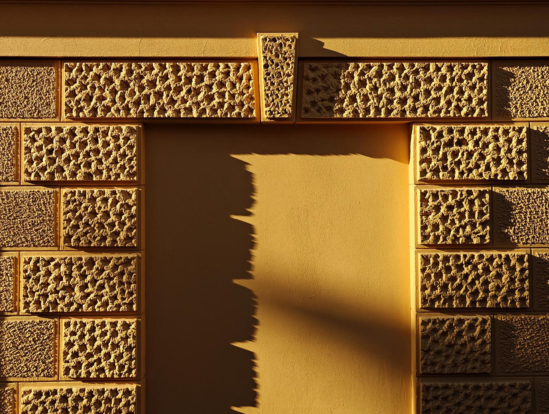 A photo showing a textured yellow stone wall with a smooth central panel where a window would be. A low bright sun to the left causes shadows in the textures and across the panel.