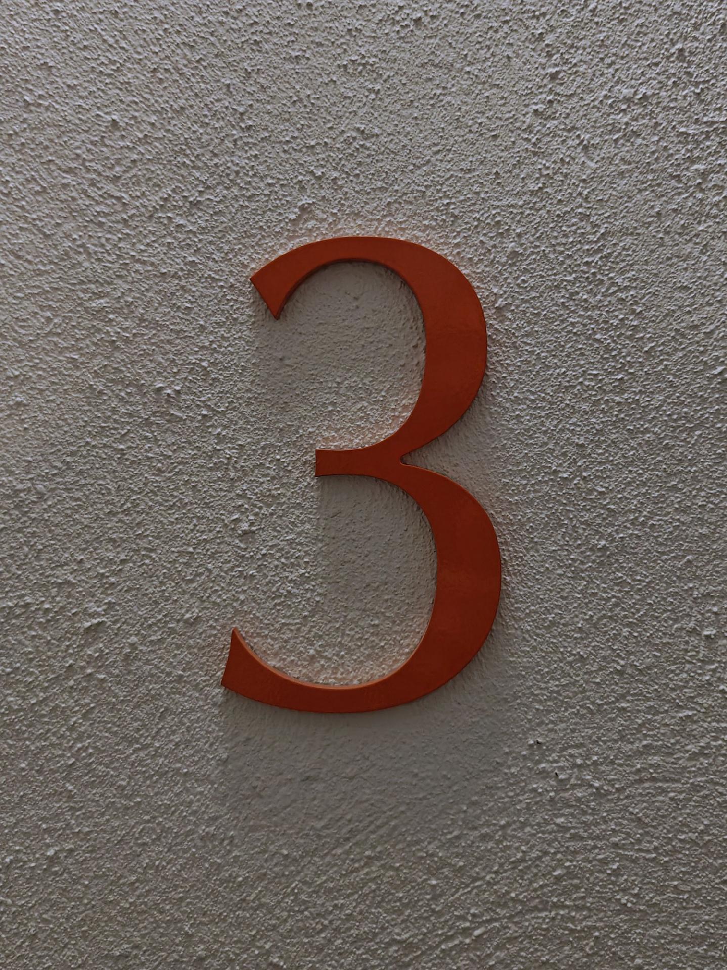 A photo of a red/orange number 3 mounted on a textured light-colored wall.