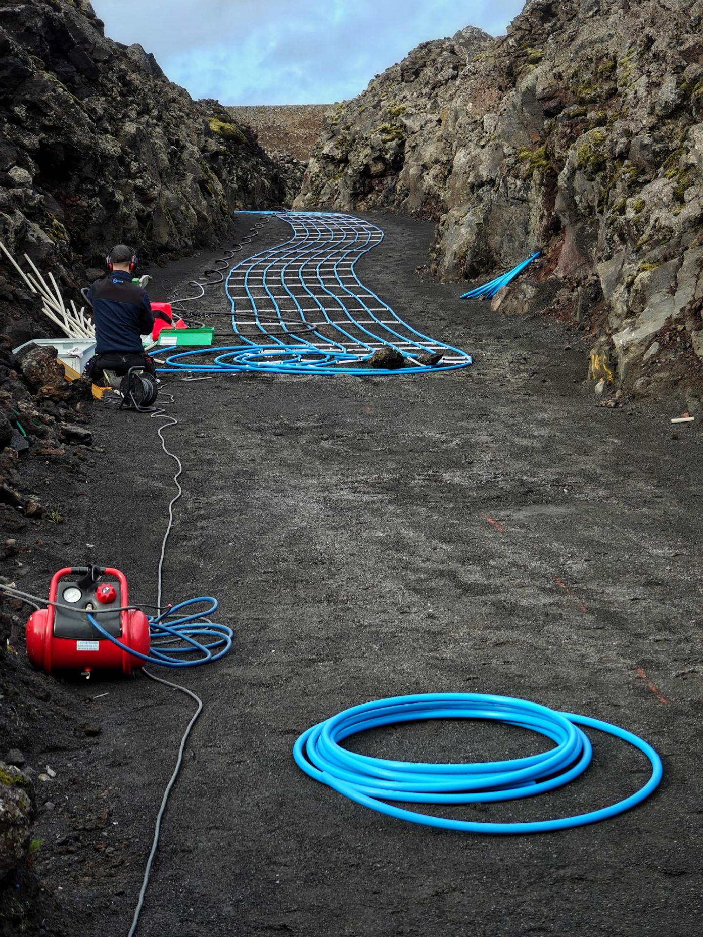 A photo of a path cut through a lage field. A person can be seen working with tools and coiled blue tubing laid across the dark rocky path between high rock walls.