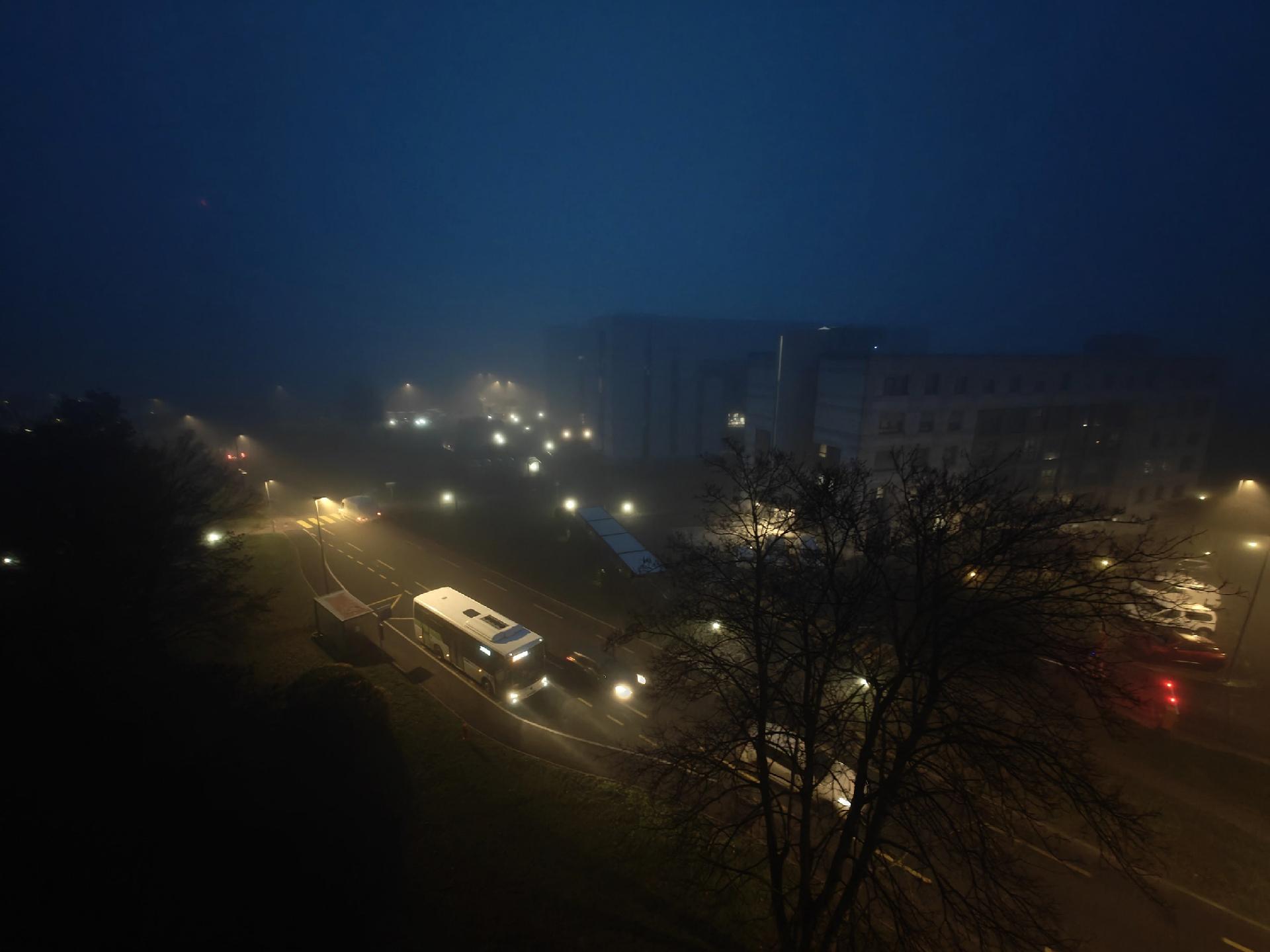 A photo looking down on foggy urban street with a bus, cars, streetlights, trees, some apartment buildings partially obscured by mist. It's early morning and still quiet dark.