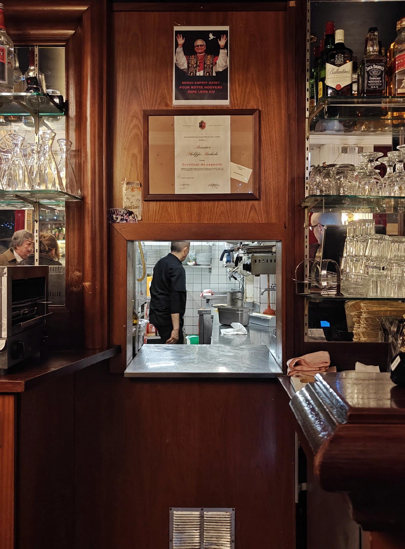 A photo showing a cozy restaurant interior with wooden shelves, bottles and glasses, and a small window revealing a cook working inside the kitchen.