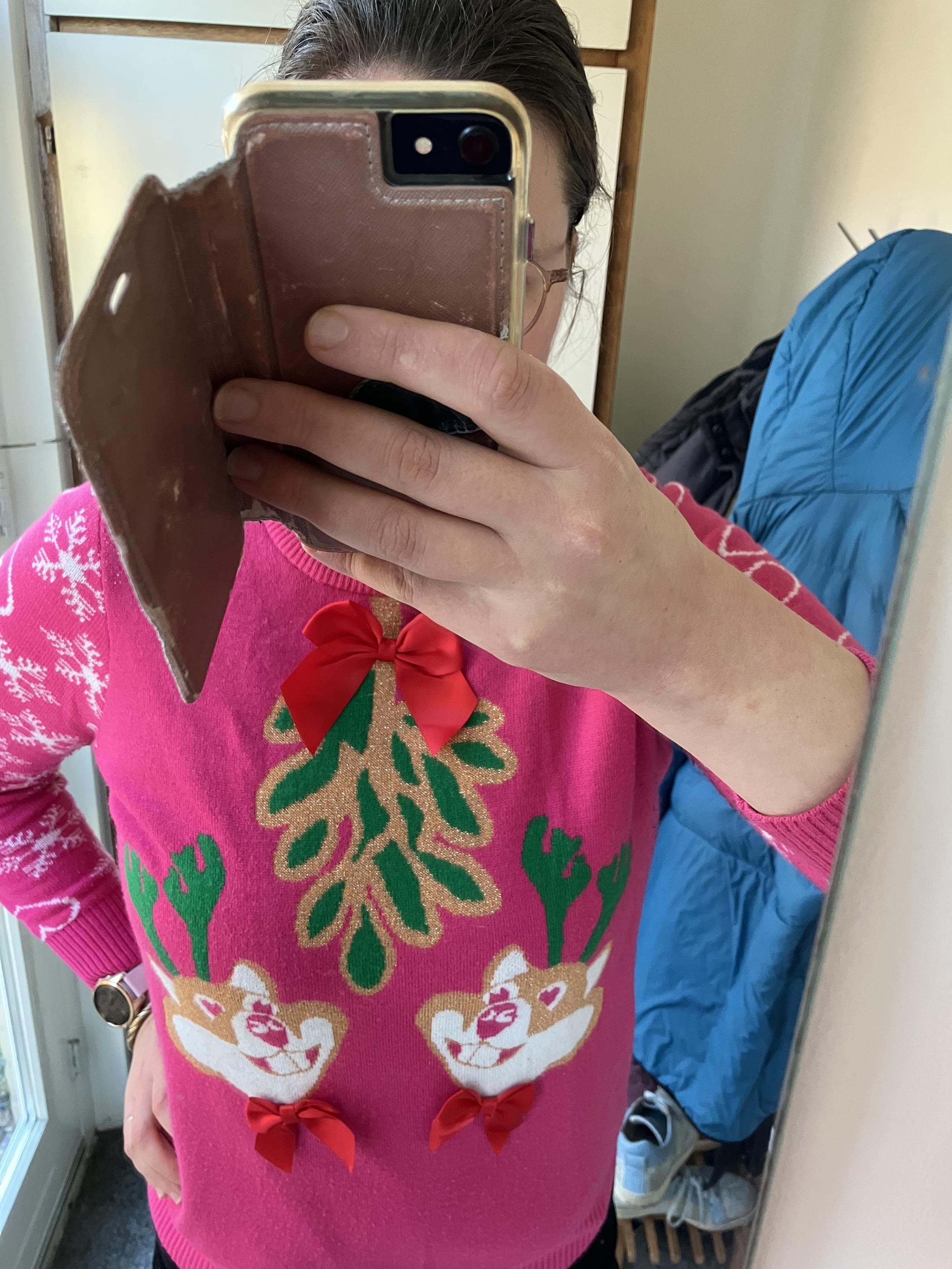 Spiegelselfie van een vrouw in een roze trui met twee hamsterhoofden met groene geweien onder een glitter maretak. Op de mouwen staan witte harten en sneeuwkristallen. De hamsters dragen opgenaaide rode satijnen strikken en de maretak is vastgeknoopt met een rode satijnen strik die daardoor ook gelijk een strik voor de drager is.