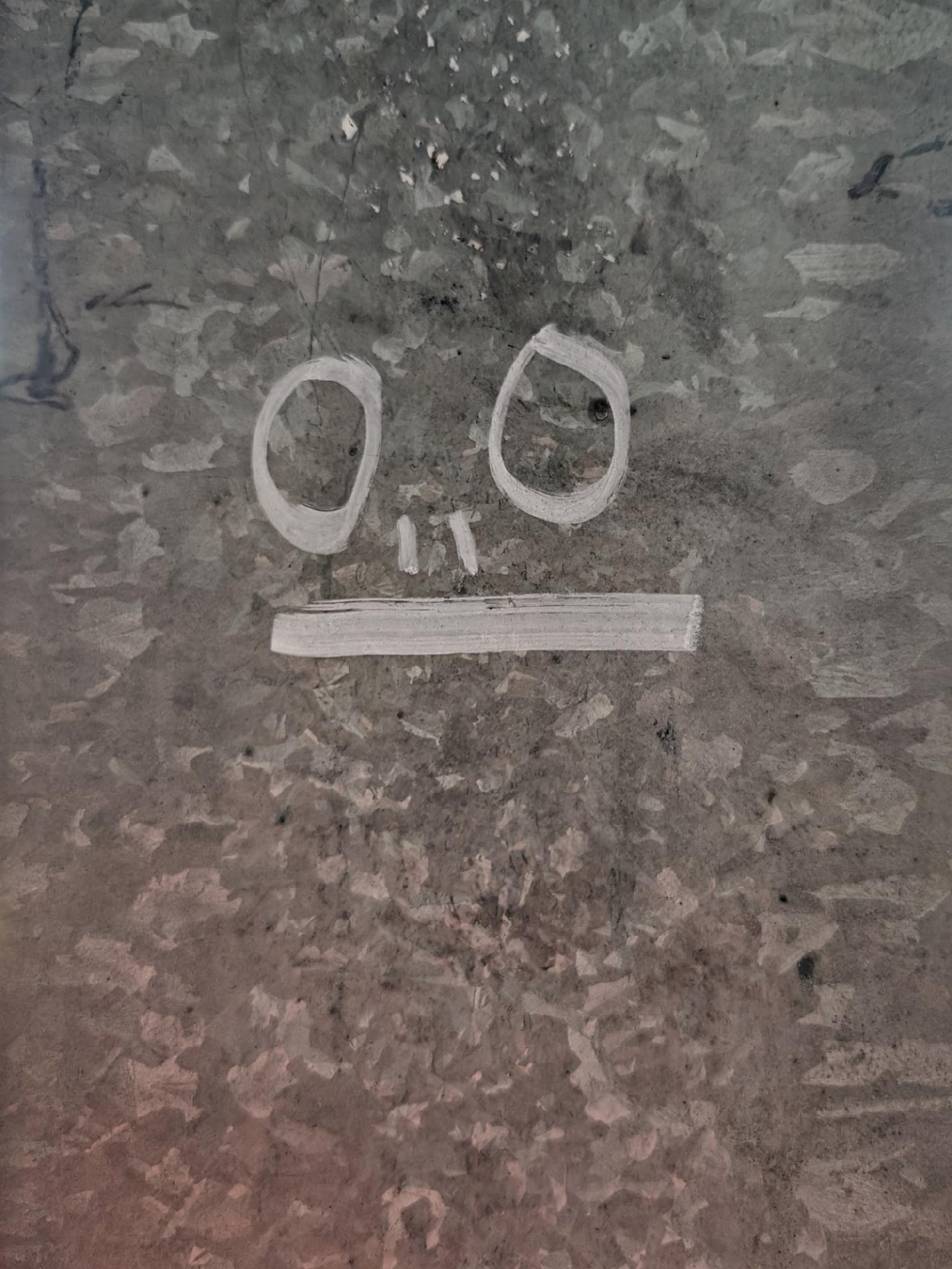 A close up photo of a grey metal pole with a simple face drawn in white paint with two oval eyes and a straight mouth.