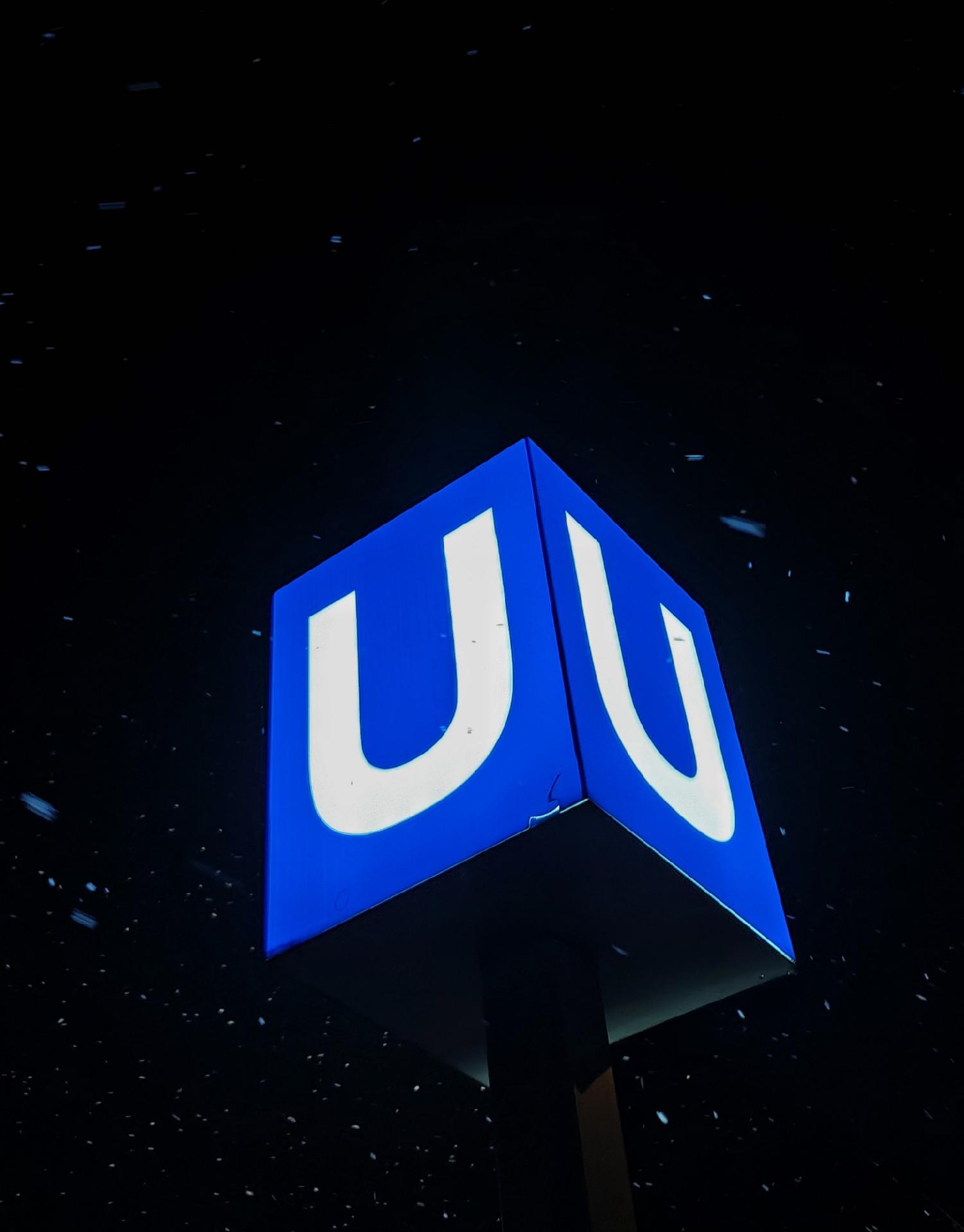 A photo of a blue illuminated subway sign with white “U”, in the dark with a light snow fall.