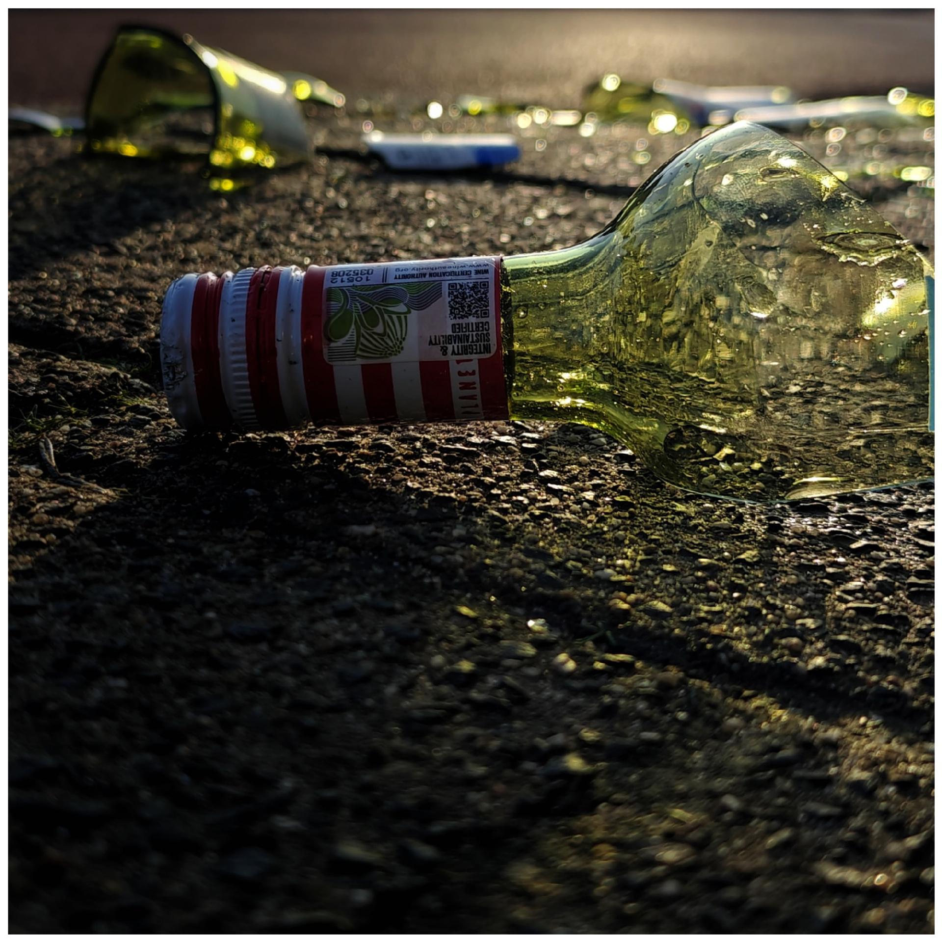 A photo showing the top of a broken glass bottle with a red and white striped label on the pavement. Pieces of greenish glass are visible in the background against low sunlight.