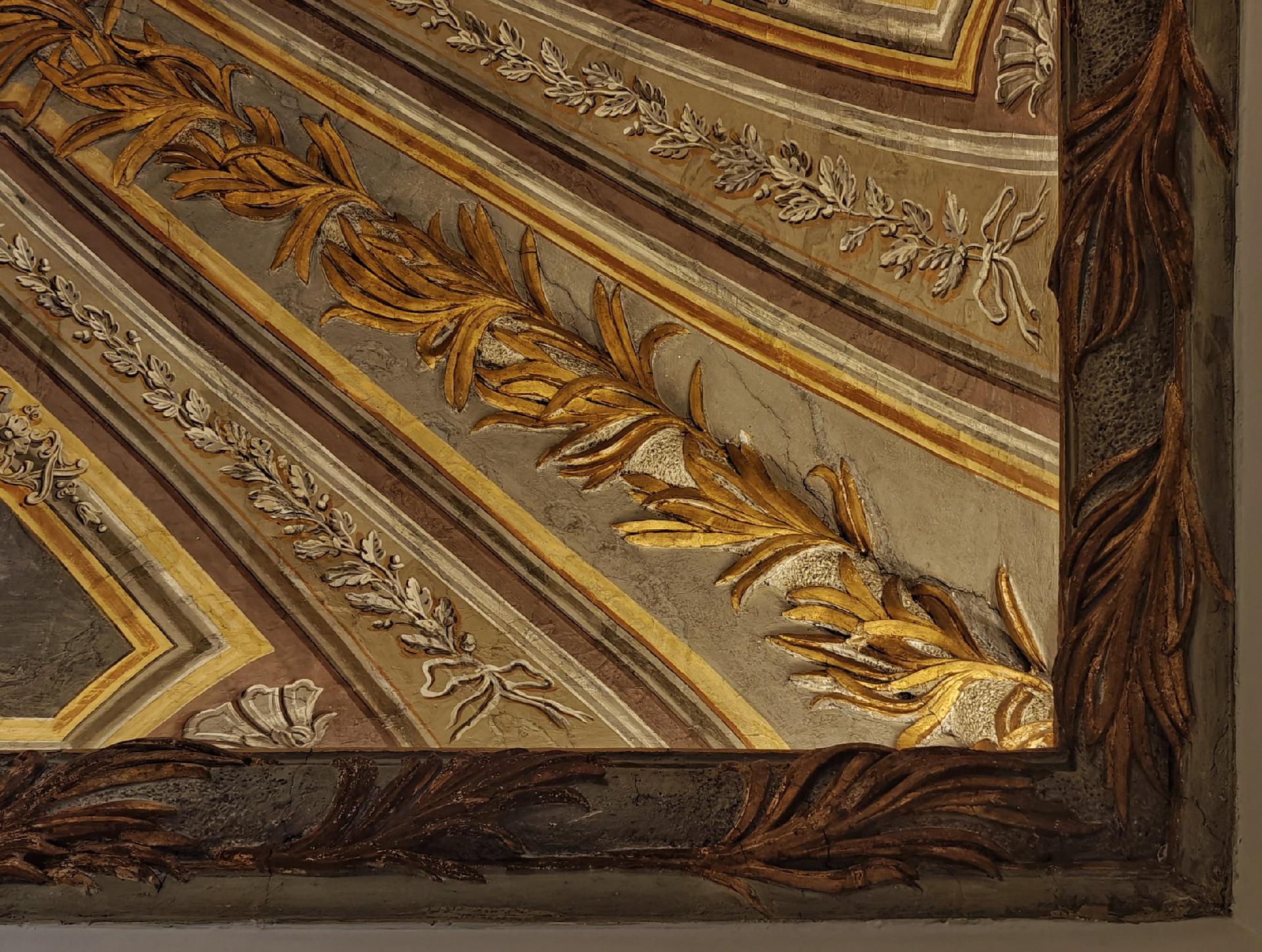 A photo of a corner of an ornate ceiling with floral and leaf patterns in gold, brown, and cream tones. The outside is dark while the rest is illuminated by hidden lights in a recess.