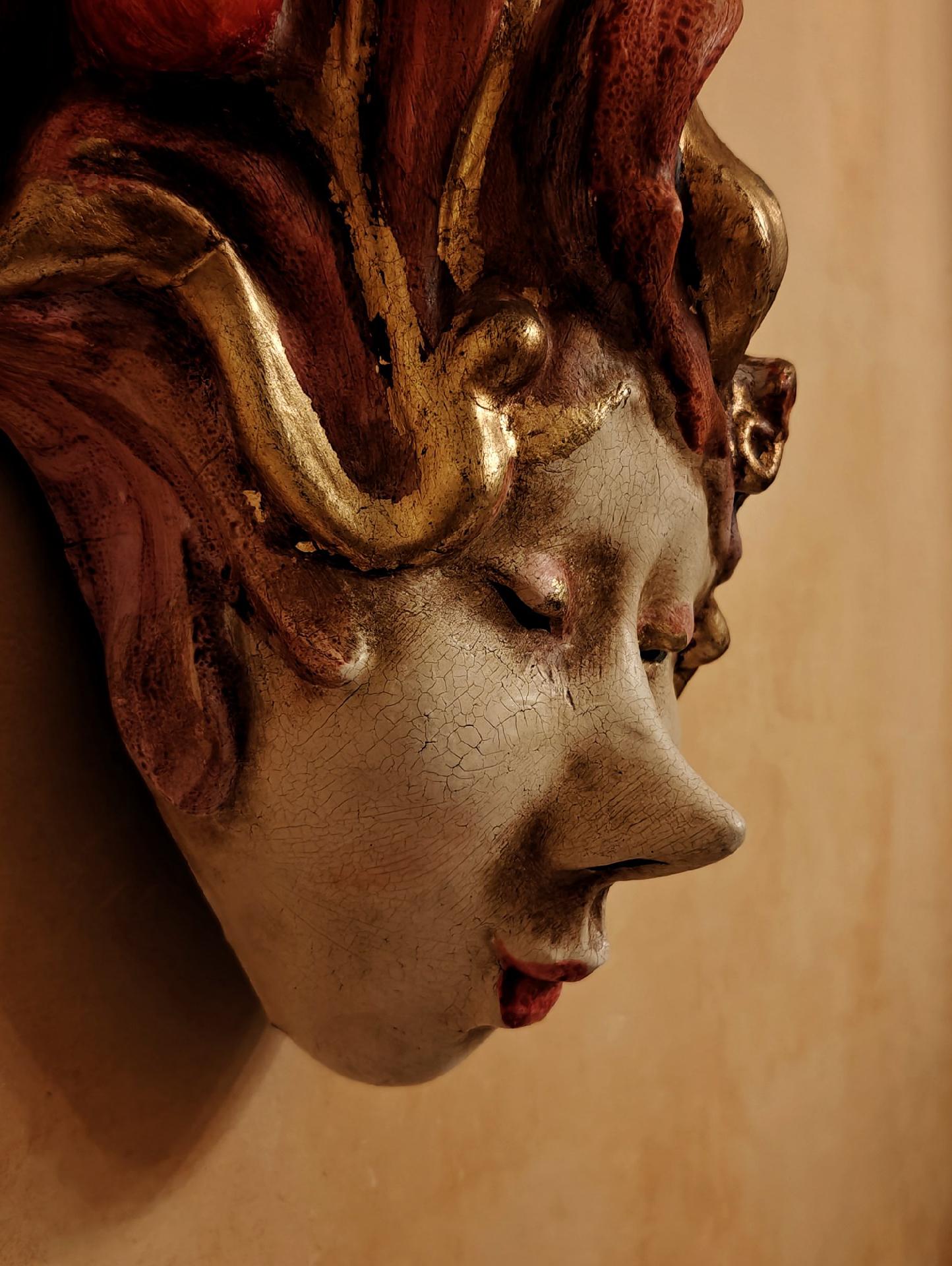 A close-up photo of a carnival mask hanging on the wall, with a pale face, red lips, and closed eyes. Cracks are visible in the painted surface.