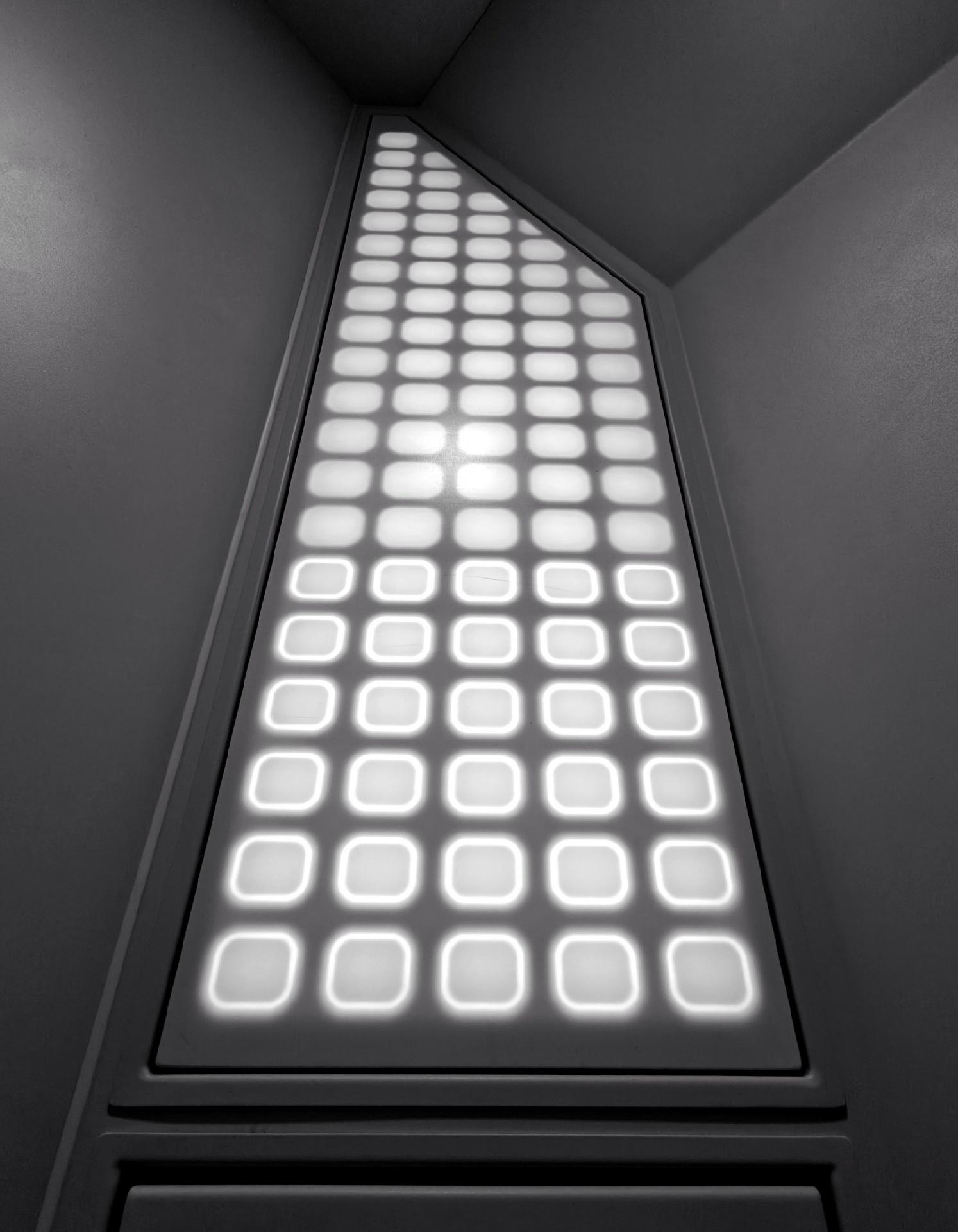 A photo showing a tall, narrow panel with a grid of glowing square lights set into a wall. The camera looks upward into a complex ceiling corner.