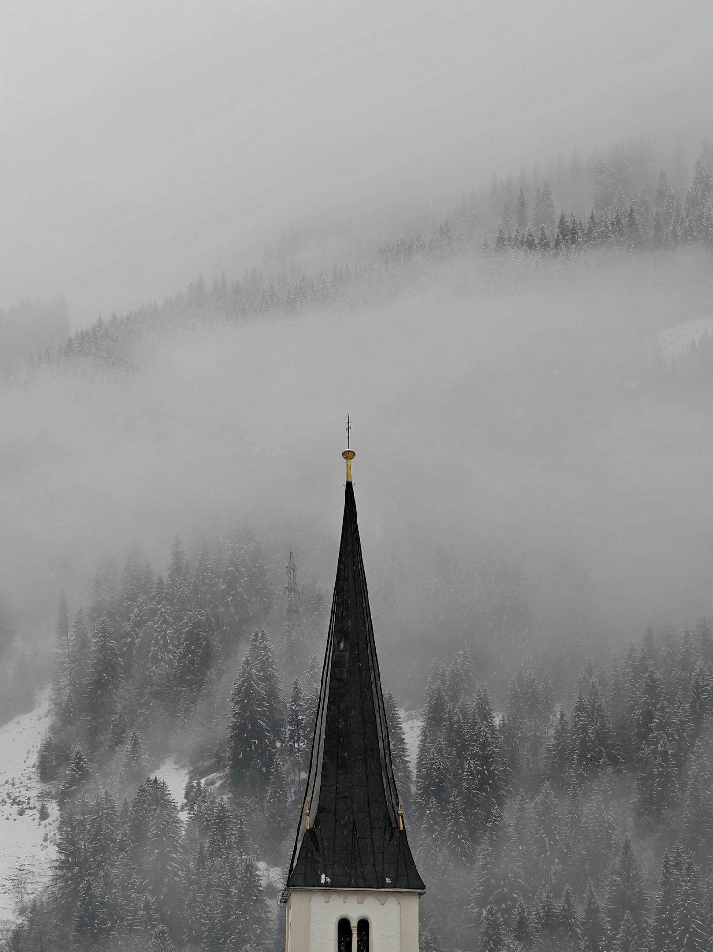 An almost black and white photo of a foggy, snow-covered hill with evergreen trees. The top of a church tower roof with a dark, pointed spire and golden top is peeking into the image from below.