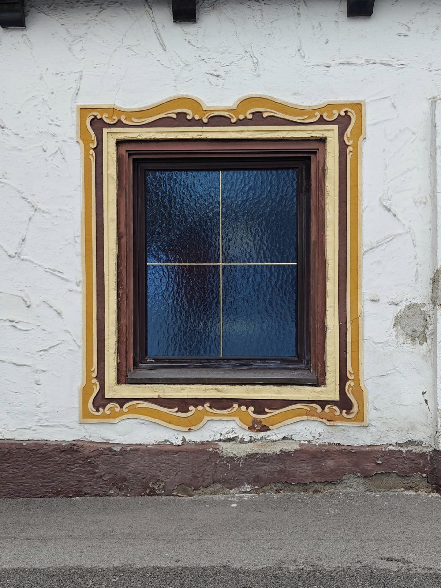 A photo of a window with blue-tinted panes, framed by ornate brown and gold plaster designs, in a white wall.