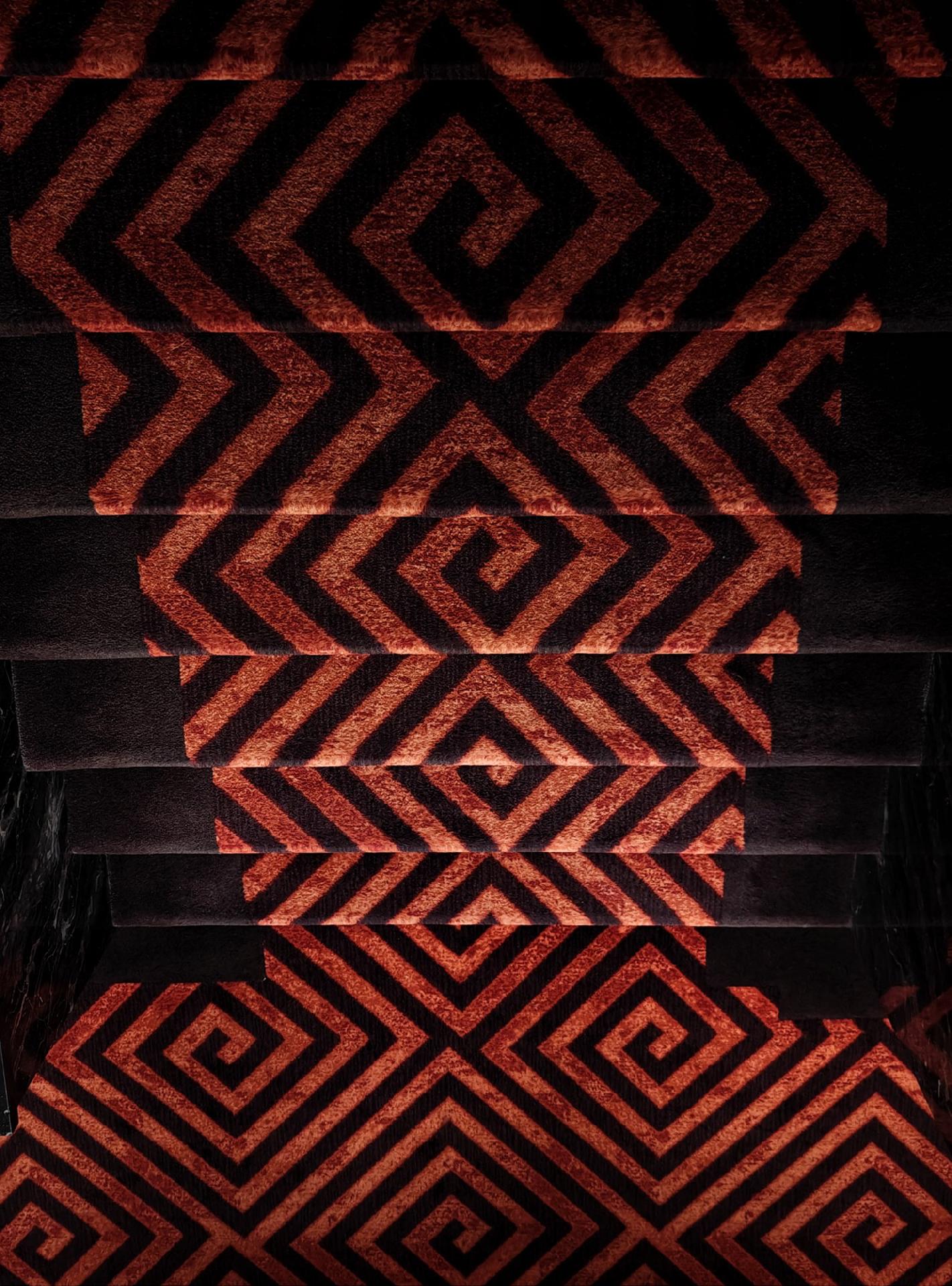 A photo looking down stairs covered by a carpet with a geometric pattern of concentric squares and diamond shapes in red and black. 