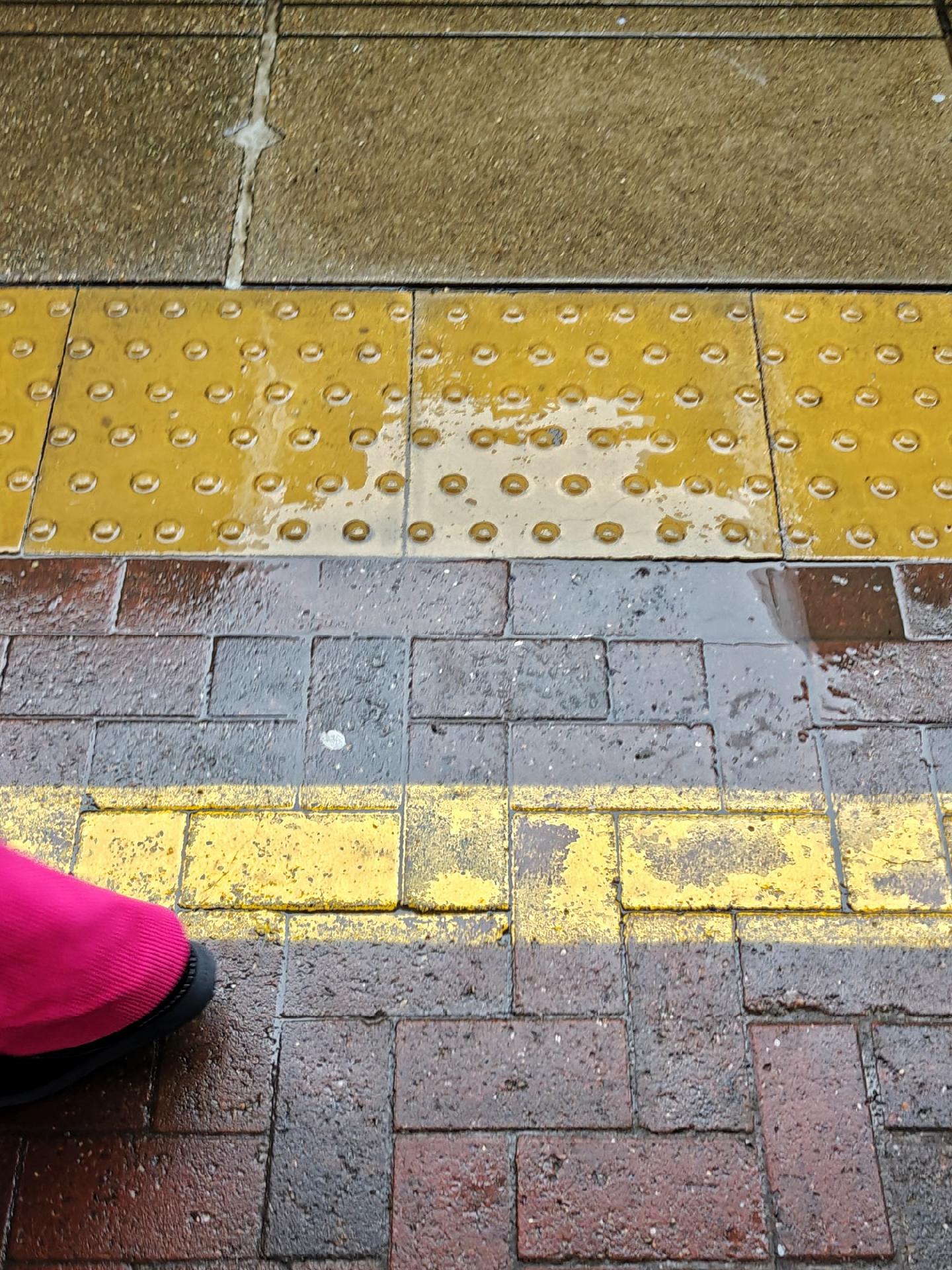 A photo looking down onto a train platform surface showing yellow tiles with raised dots at top, gray bricks, and a yellow painted line. A small part of leg with bright magenta fabric is just leaving the view at the bottom left corner.