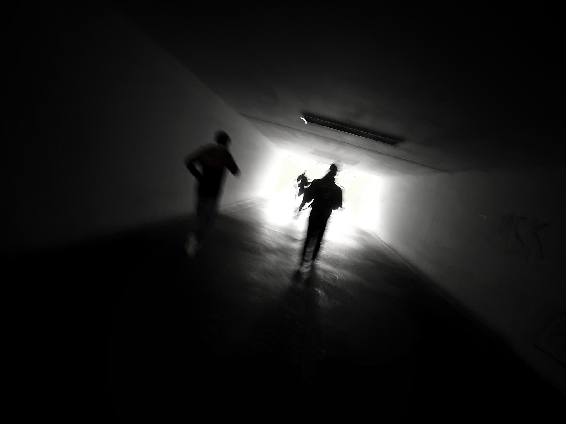 A black and white photo with motion blur showing silhouetted figures in a dark space moving toward or bright light coming from an opening.