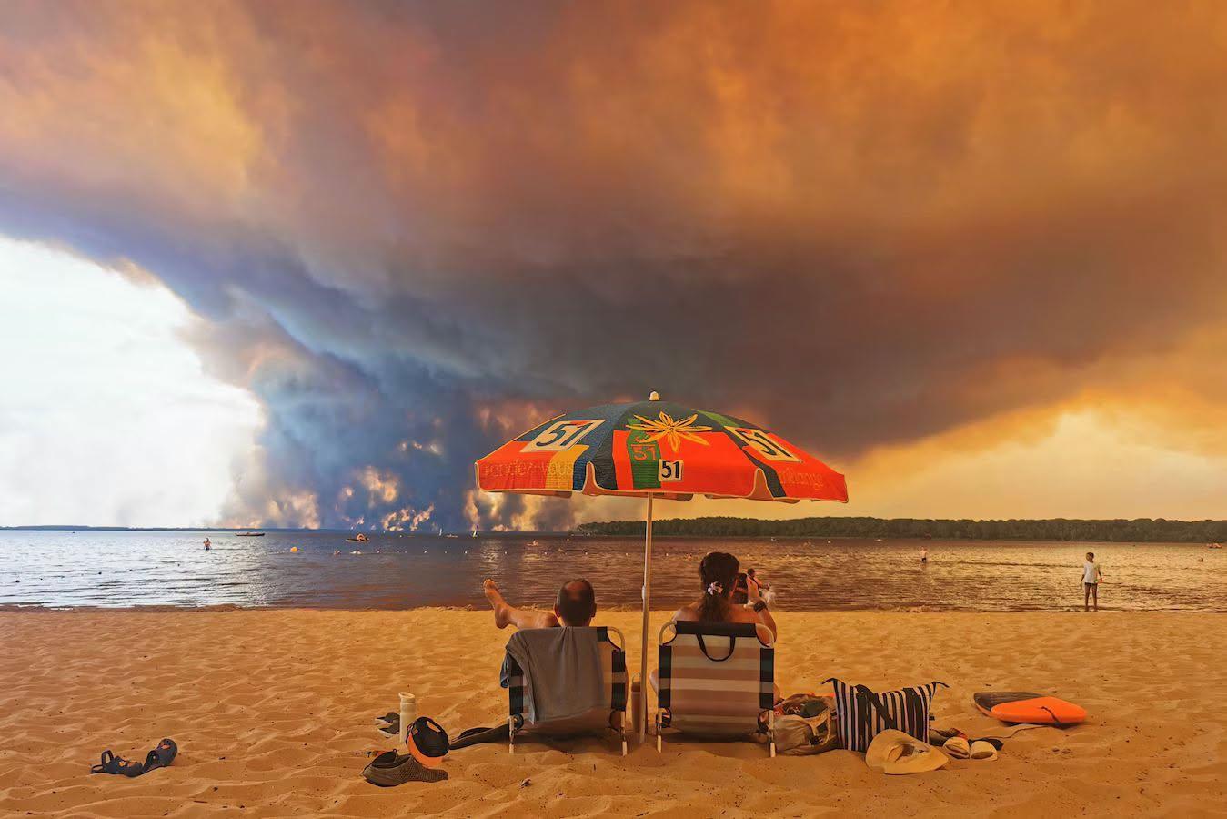 Een stel zit aan de kust van Frankrijk aan zee onder een parasol. Een oranje gloed en rook van de bosbranden daar trekt over zee.