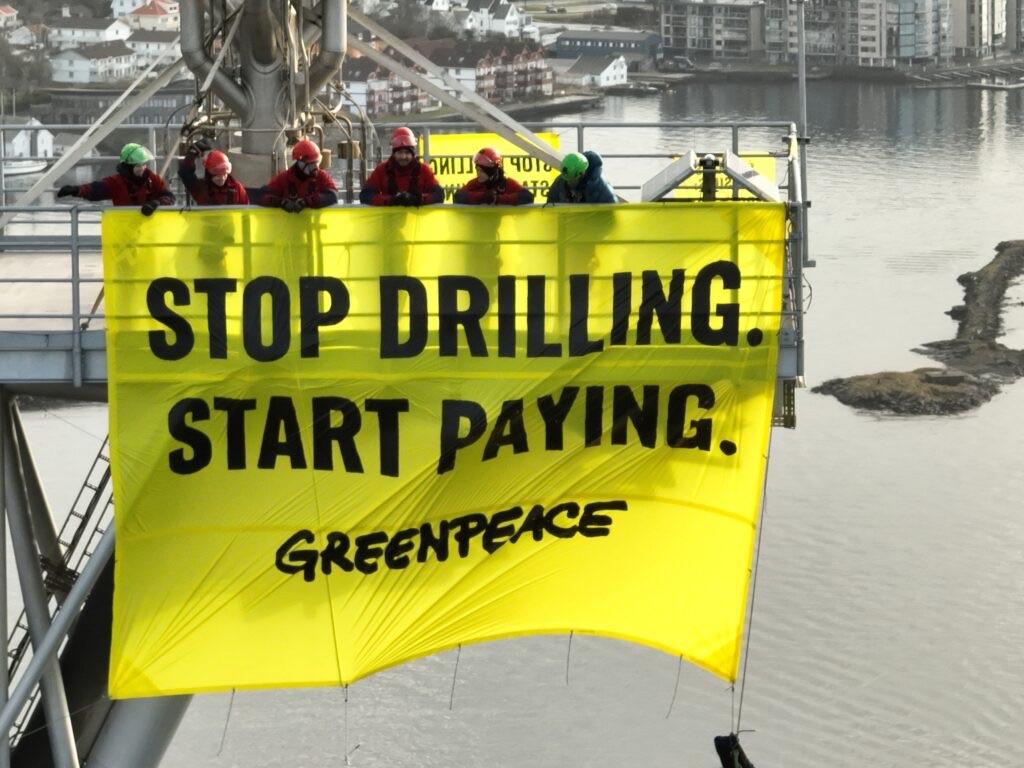 Foto van demonstranten van Greenpeace met een vlag waarop staat “Stop drilling, start paying”.