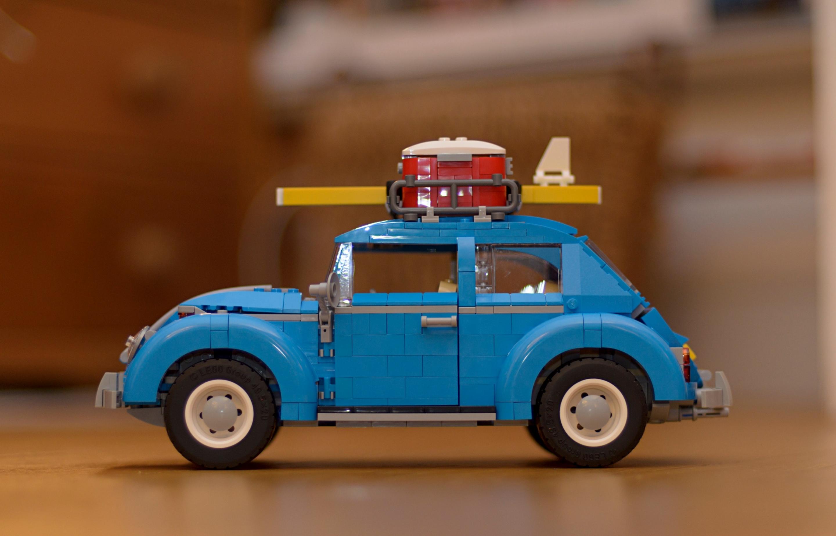 A photo of a blue LEGO Volkswagen Beetle car with a suitcase and surfboard mounted on the roof. The model stands on a wooden surface, with a blurred background.