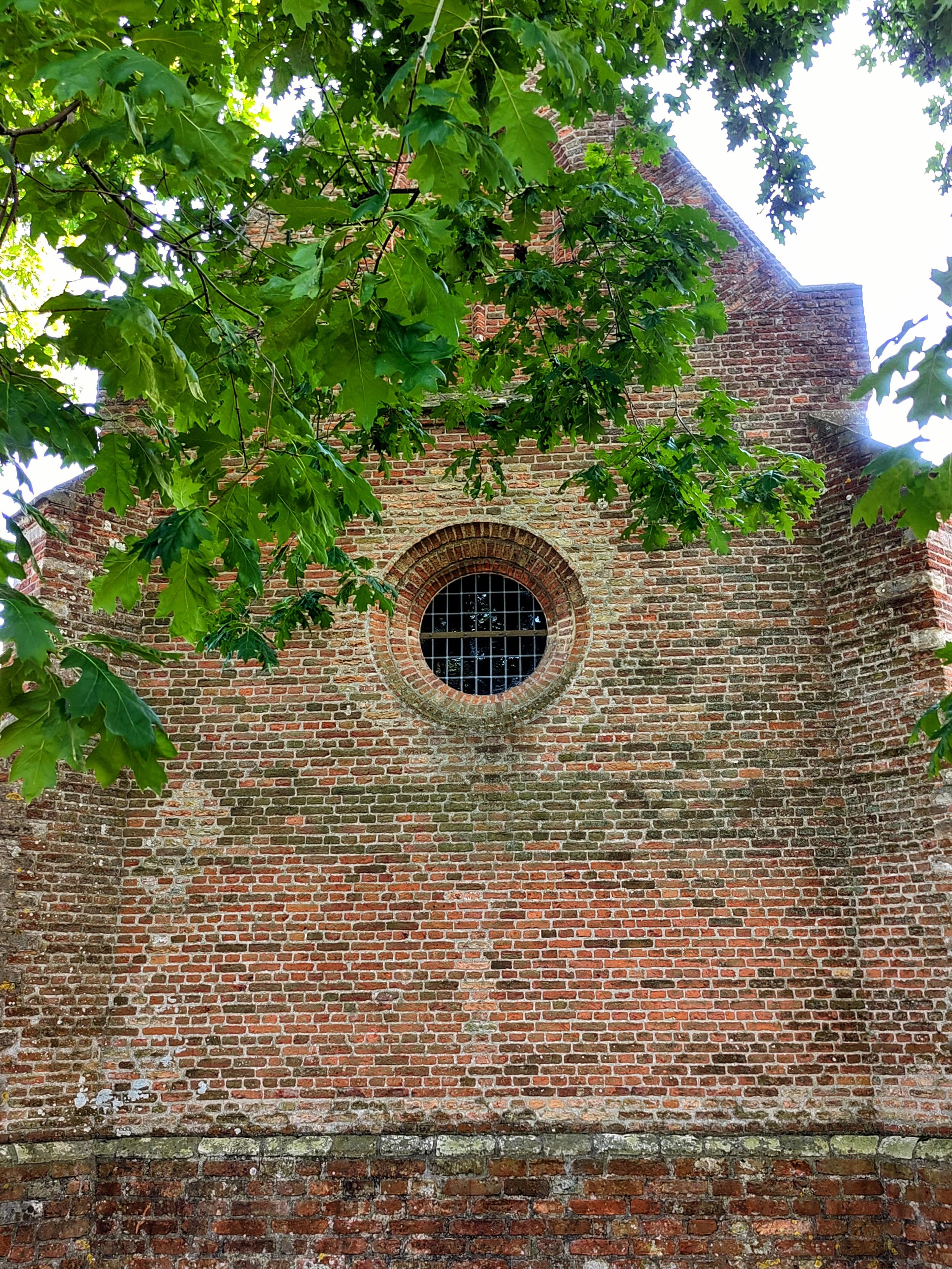 Bakstenen geven van een kerk met een rond raam onder een groene bladeren van een loofboom ¦ Brick facade of a church with a round window below green foilage of a tree