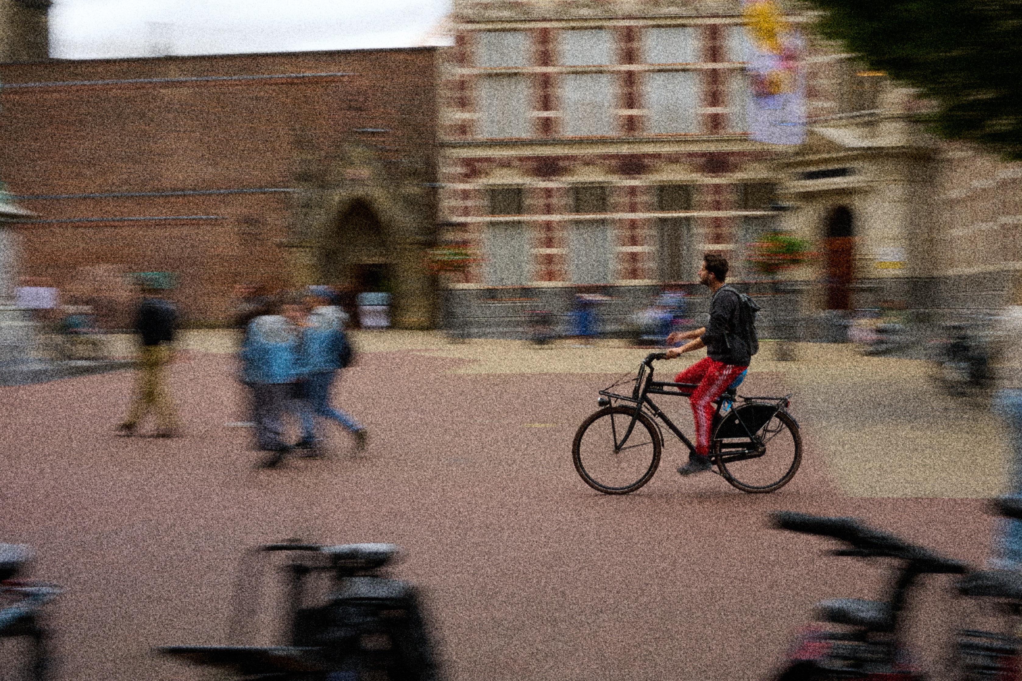 A person rides a bicycle through a busy square with blurred pedestrians and historical buildings in the background. The cyclist wears a grey shirt and red pants, while multiple people walk nearby, creating a lively urban atmosphere.