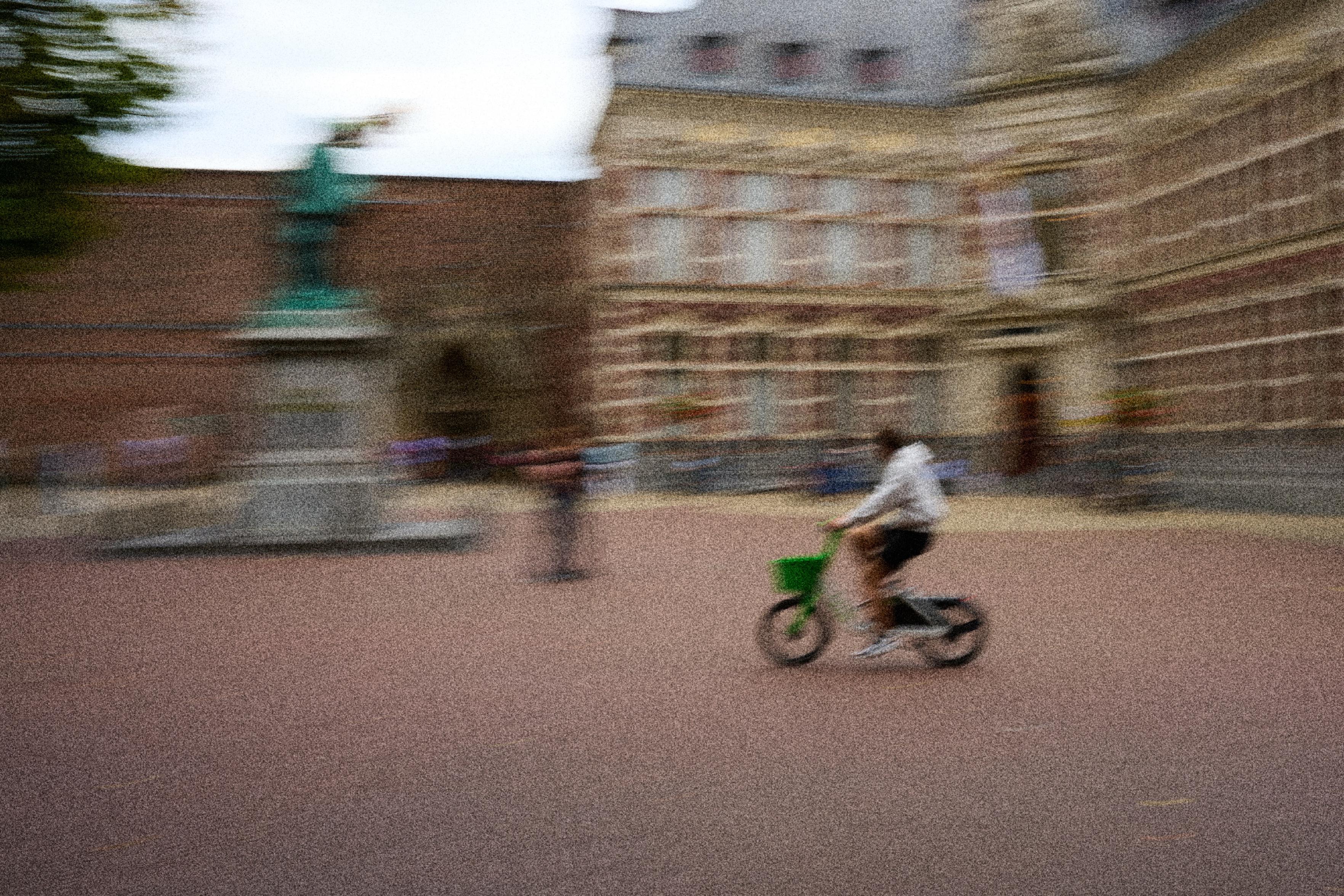 A blurred image of a person riding a bicycle with a green basket in a plaza, with a statue in the background and several people walking. The scene conveys a sense of movement and activity.