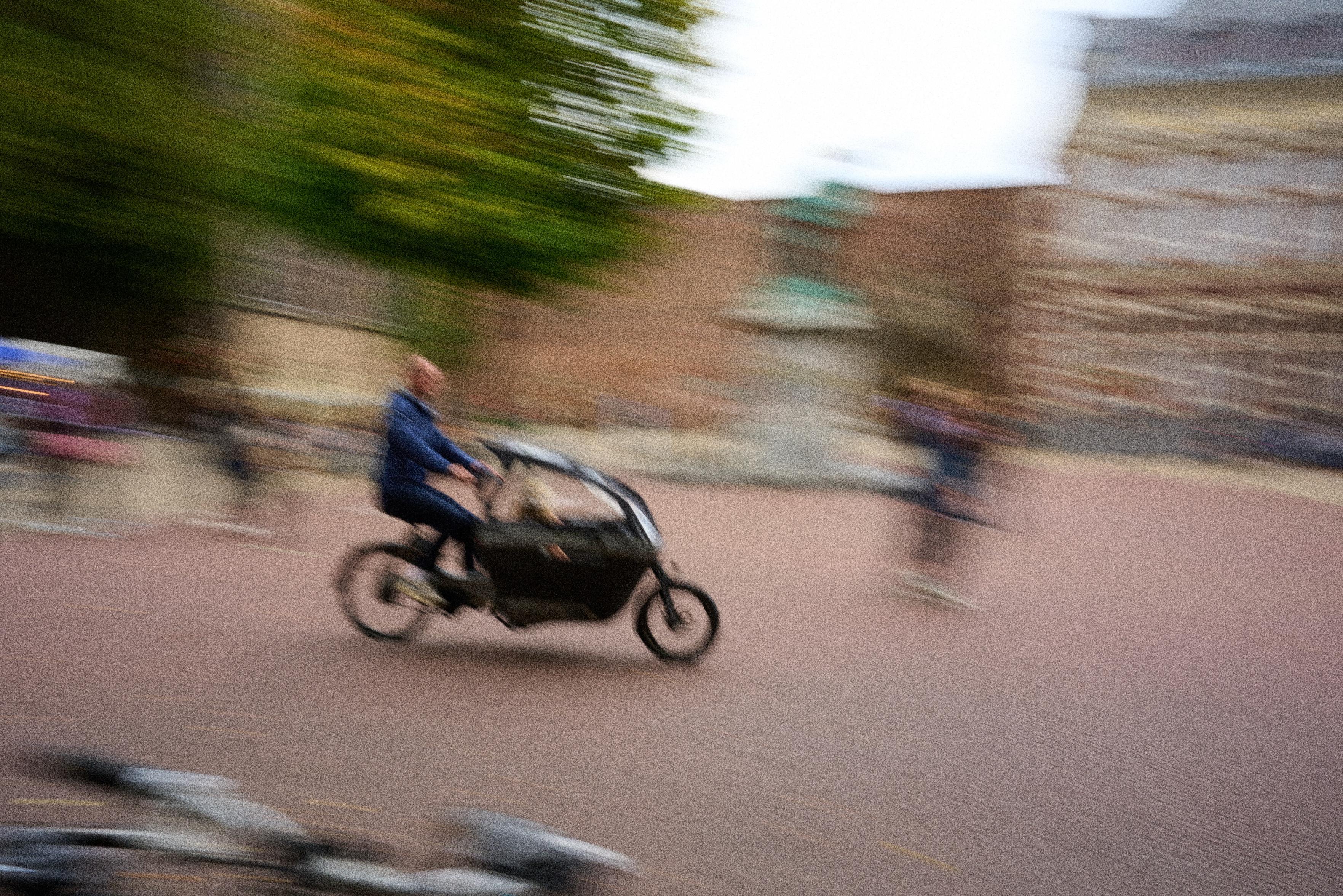 A blurred image of a person riding a cargo bike through a busy outdoor area, with trees and buildings in the background. Other people are present, adding to the sense of motion and activity.