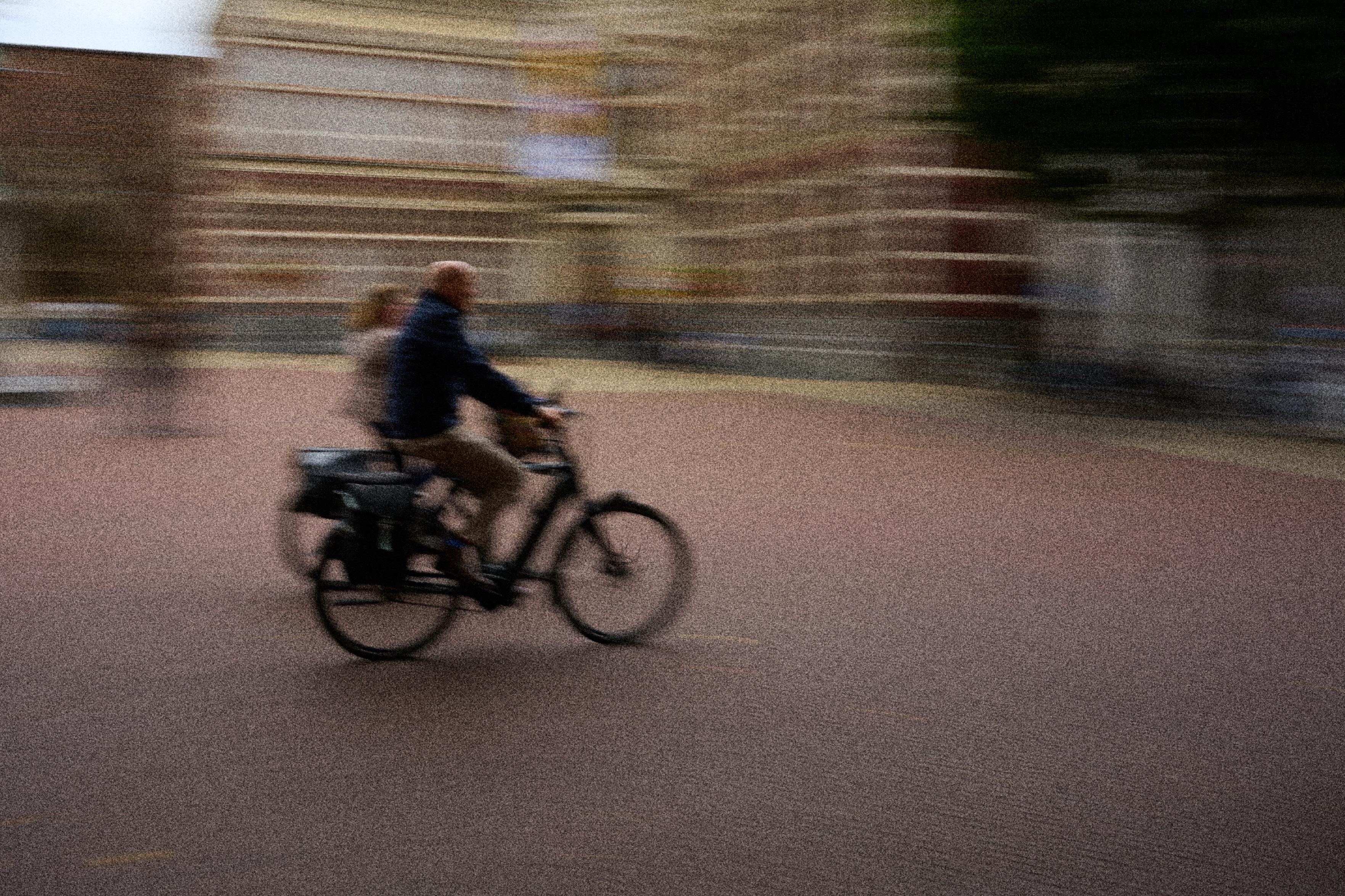 A cyclist and a passenger are moving quickly on a bicycle in a blurred city setting, with buildings and a sidewalk in the background.